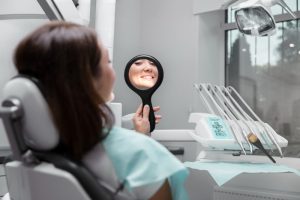 Patient in a dental chair holding a handheld mirror and smiling at her reflection in a clinical setting