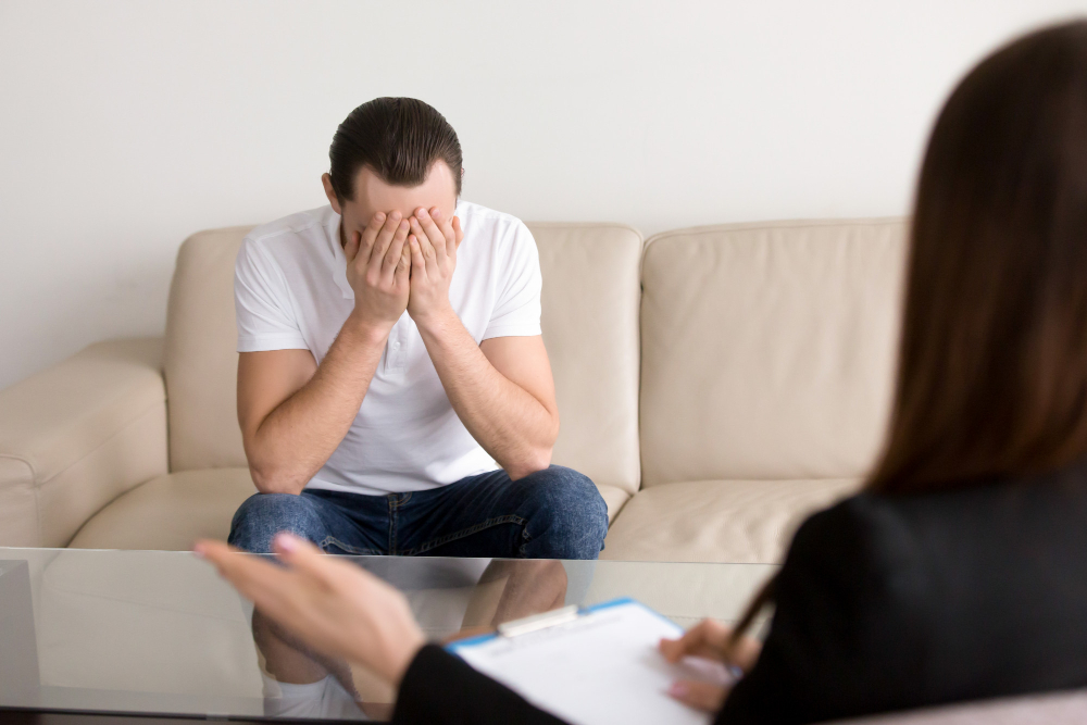 Man sits on a beige sofa, hands covering his face in distress during a therapy session at home, with a clinician taking notes nearby in the foreground.] }]}Oops I messed again. I should output clean. Let's do final correctly.