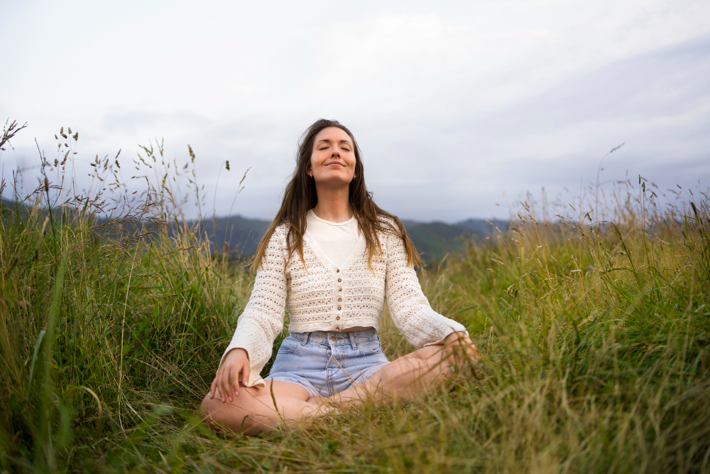 Woman sits cross‑legged in a grassy field with eyes closed, enjoying a peaceful moment outdoors, with rolling hills in the distance.