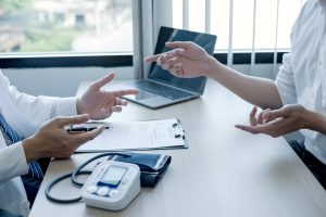 Two professionals discuss a medical chart at a desk, with a laptop and blood pressure monitor nearby.