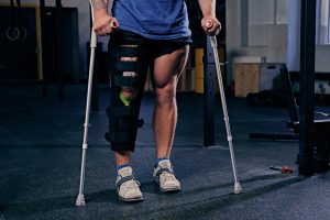 Athlete standing with crutches and a knee brace on the left leg in a gym setting.