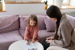 A young girl draws with a marker on a clipboard while an adult sits beside her on a pink sofa in a bright living room.