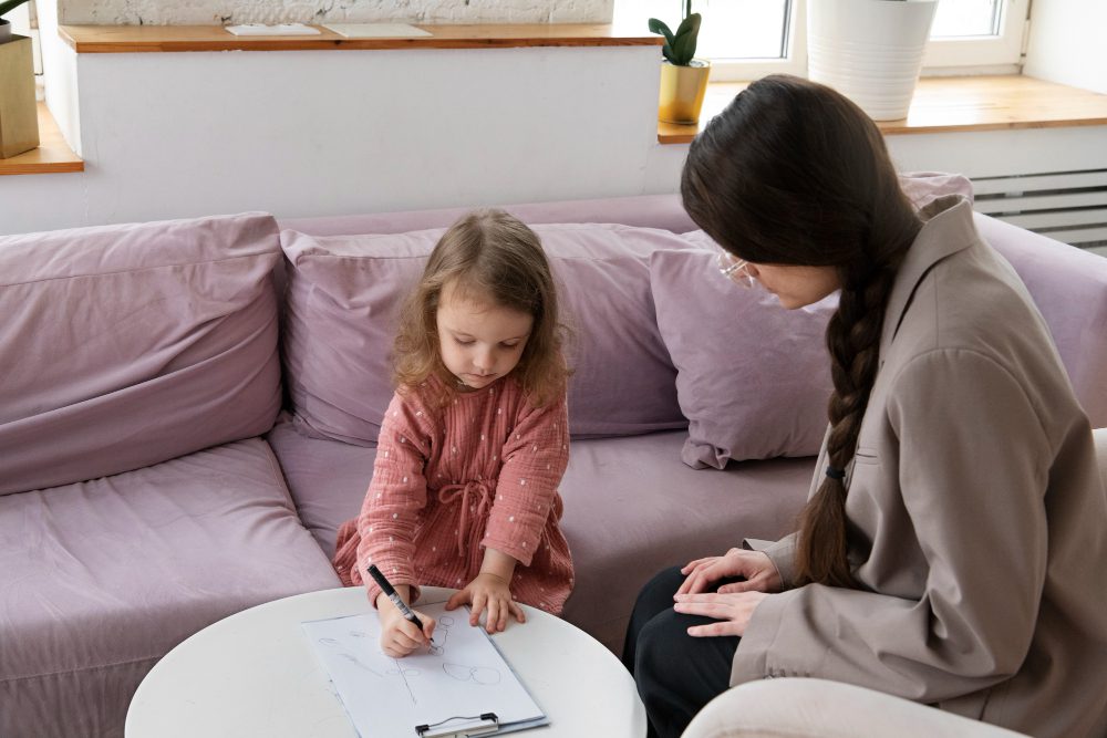 A young girl draws with a marker on a clipboard while an adult sits beside her on a pink sofa in a bright living room.