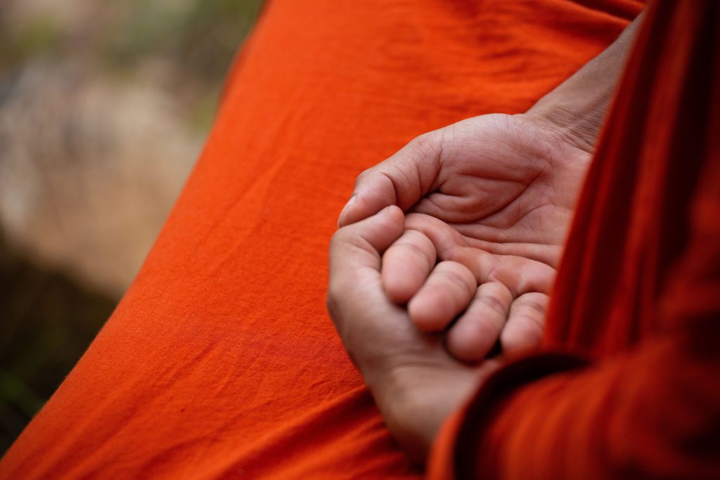 Close-up of cupped hands resting on a bright orange robe, suggesting meditation or prayer.