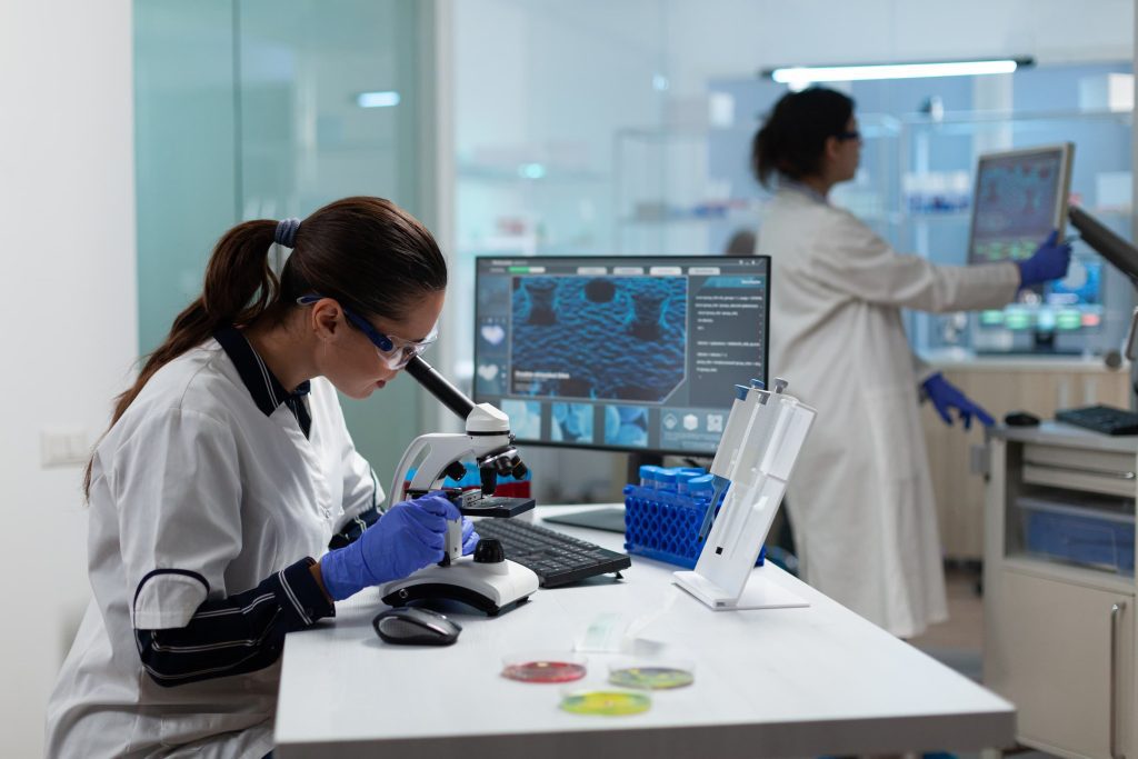 Female scientist wearing safety goggles and gloves looks into a microscope at a lab bench, with a computer monitor nearby.